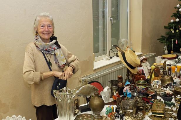 Eine ältere Frau steht lächelnd an einem Tisch mit verschiedenen Antiquitäten und Dekorationsgegenständen.