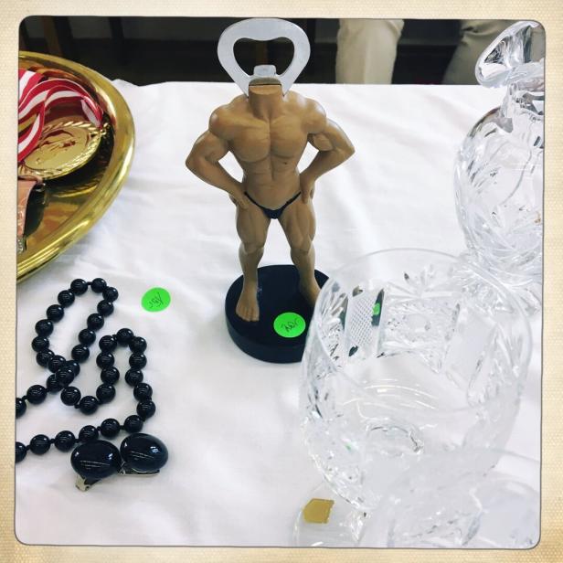 Ein Flaschenöffner in Form eines muskulösen Männerkörpers steht zwischen Kristallgläsern und einer schwarzen Perlenkette.