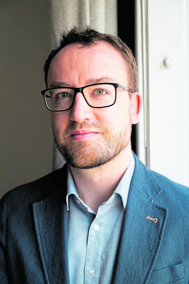 Mann mit Brille, Bart und kurzem Haar trägt ein blaues Sakko und steht vor einem Fenster mit hellem Hintergrund.