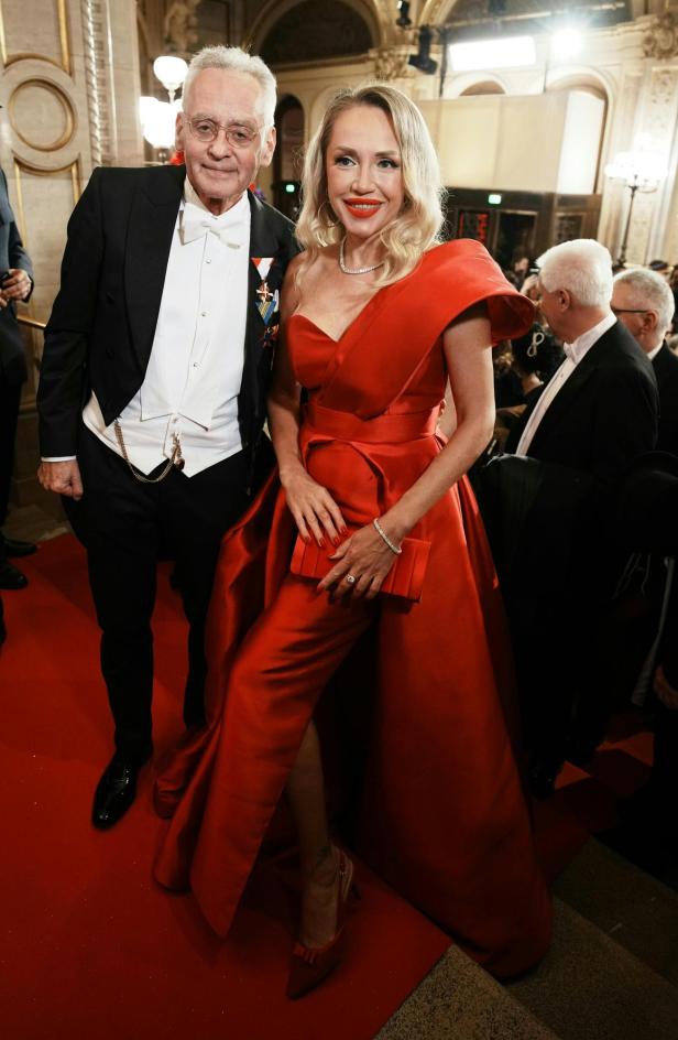 Ein elegant gekleidetes Paar steht auf einem roten Teppich in einem prunkvollen Saal, umgeben von weiteren festlich gekleideten Gästen.