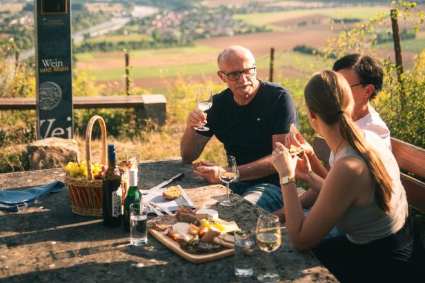 Drei Personen sitzen an einem Steintisch im Freien mit Weinflaschen, Gläsern und einer Käseplatte vor einer Landschaft mit Weinbergen.