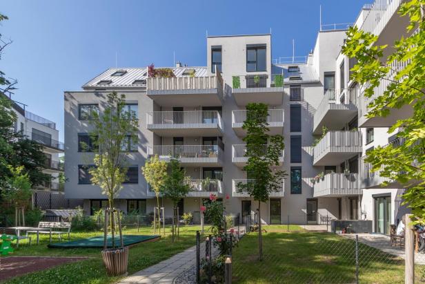 Mehrstöckiges modernes Wohnhaus mit Balkonen, davor ein grüner Garten mit Bäumen, Sträuchern und Sitzbänken.