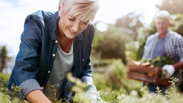 Frau arbeitet lächelnd im Garten, im Hintergrund steht ein Mann mit einer Holzkiste voller Gemüse.