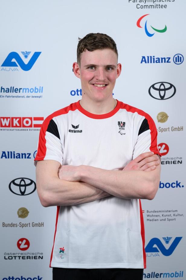 Johannes Aigner mit verschränkten Armen trägt ein Sporttrikot mit österreichischem Wappen vor Logos.