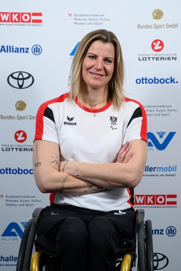 Christina Obwexer hat die Arme verschränkte und trägt ein österreichisches Sporttrikot vor einer Wand mit Sponsorenlogos.
