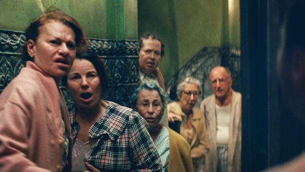 Sechs ältere Menschen stehen in einem Flur und blicken erschrocken und überrascht in eine Richtung.