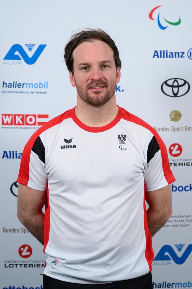 Thomas Grochar, ein Mann mit kurzem Bart, trägt ein weiß-rotes Sporttrikot mit österreichischem Wappen und Paralympics-Logo, steht vor Sponsorenlogos.