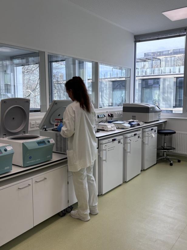 Eine Person in weißer Kleidung arbeitet an Laborgeräten neben mehreren Kühlschränken in einem hellen Raum mit großen Fenstern.