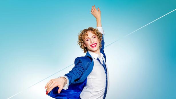 Person mit lockigen Haaren, blauem Anzug und Make-up lächelt und streckt die Arme vor blauem Hintergrund aus.