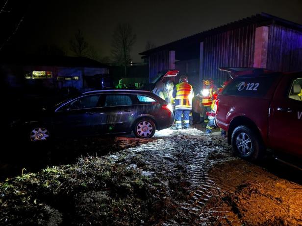 Mehrere Feuerwehrleute in Warnwesten stehen nachts bei zwei Autos auf schlammigem Boden vor einem Gebäude.