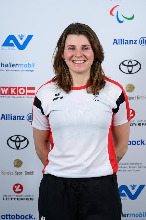  Elisabeth Aigne, eine junge Frau mit schulterlangem braunem Haar trägt ein weißes Sporttrikot mit roten Akzenten und lächelt vor Sponsorenlogos.