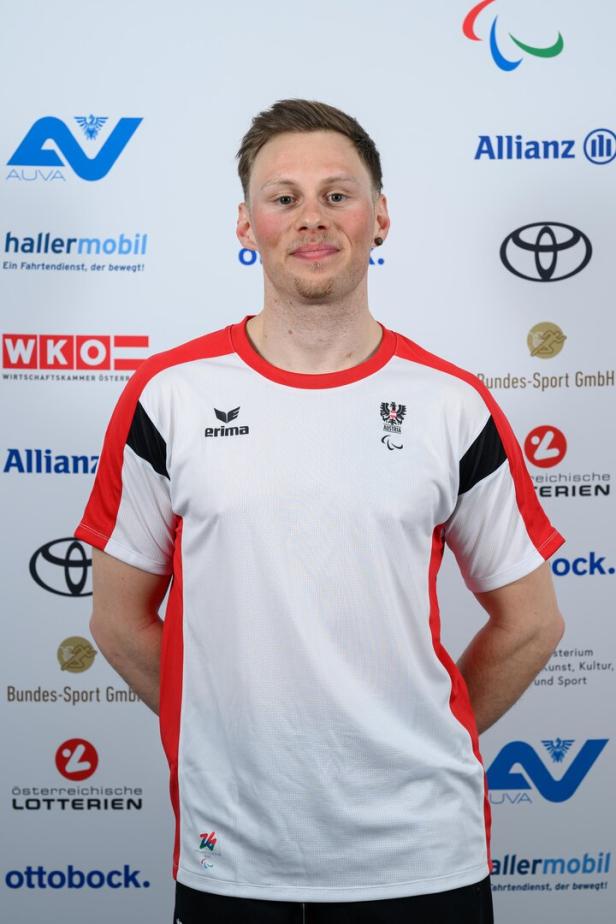 Stefan Egger-Riedmüller im weißen Sporttrikot mit roten Akzenten steht vor einer Sponsorenwand und lächelt leicht.