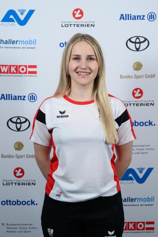 Elina Stary trägt ein weiß-rotes Sporttrikot und steht vor einer Sponsorenwand.