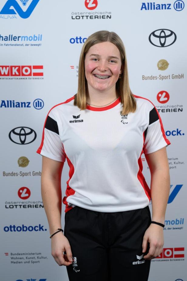 Lilly Sammer trägt Sportkleidung mit Österreich-Logo und lächelt vor Sponsorenwand.