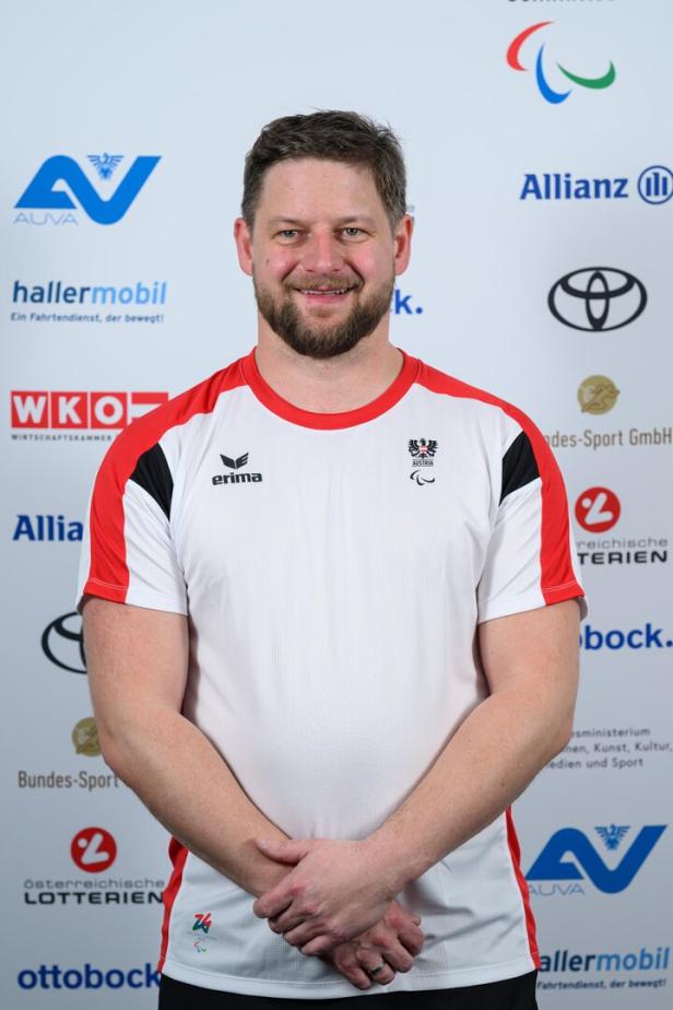 Michael Scharnagl trägt ein weißes Sporttrikot mit roten Akzenten und steht vor einer Sponsorenwand.