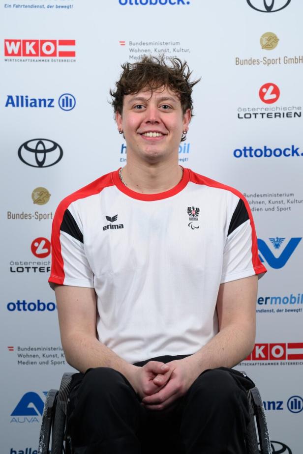 Sebastian Rigby lächelt und trägt ein Sporttrikot mit Österreich-Logo vor Sponsorenlogos.