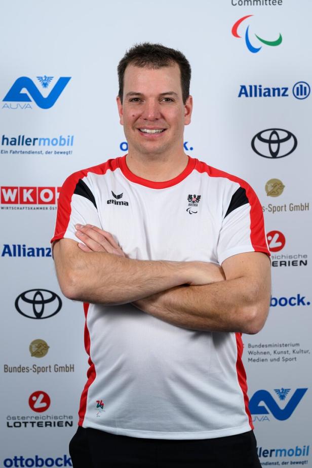 Markus Salcher im weißen Sportshirt mit roten Akzenten steht mit verschränkten Armen vor einer Sponsorenwand und lächelt.