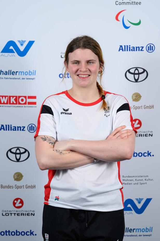 Veronika Aigner steht lächelnd mit verschränkten Armen vor einer Sponsorenwand.