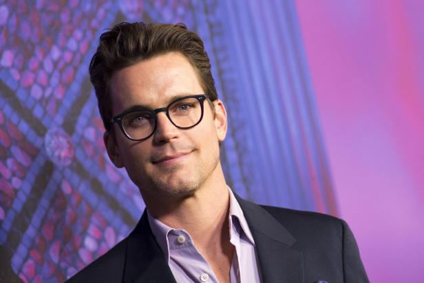 Matt Bomer trägt ein helles Hemd, ein dunkles Sakko und eine Brille vor einem violetten Hintergrund.