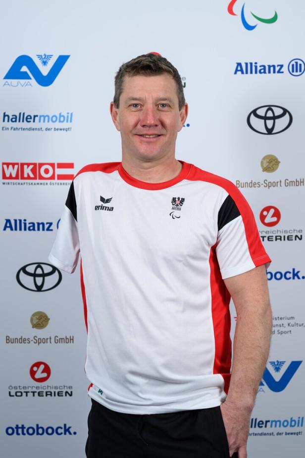 Oliver Dreier in einem weiß-roten Sporttrikot mit österreichischem Wappen steht vor einer Sponsorenwand und lächelt.