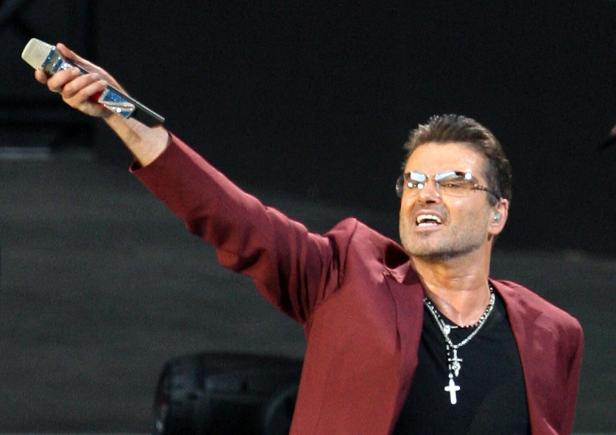 George Michael singt in einem weinroten Sakko mit ausgestrecktem Arm und Mikrofon vor einem dunklen Hintergrund.