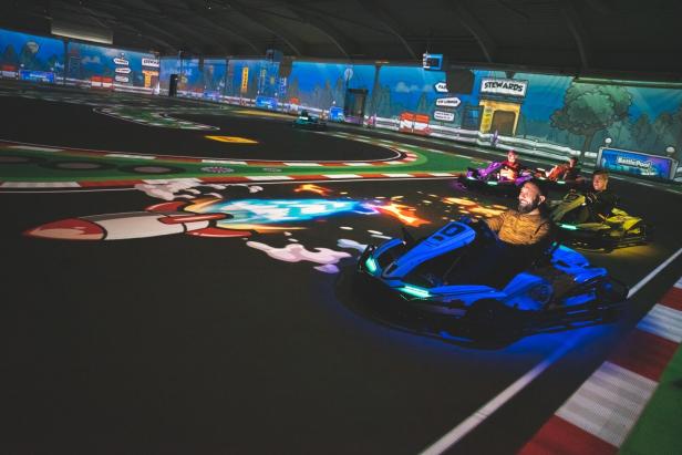 Mehrere Personen fahren beleuchtete E-Karts auf einer bunten Indoor-Strecke mit projizierten Animationen und Comic-Grafiken.