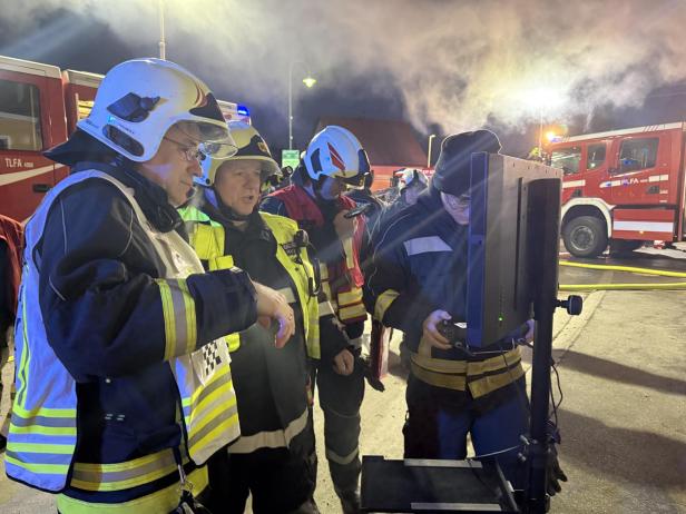 Vier Feuerwehrleute in Einsatzkleidung stehen vor einem Monitor, im Hintergrund sind Feuerwehrfahrzeuge und Rauch zu sehen.