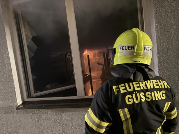 Ein Feuerwehrmann blickt durch ein Fenster in einen verrauchten Raum mit sichtbaren Flammen im Inneren.