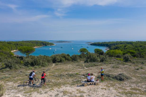 Radfahrer und Wanderer genießen den Ausblick auf eine Bucht mit Segelbooten und grüner Küste.