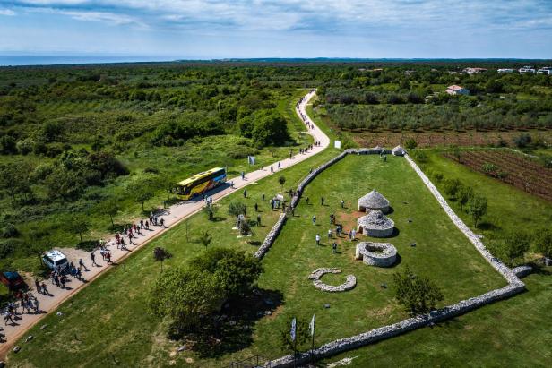 Menschen spazieren auf einem Weg neben einem Bus und traditionellen Steinhütten in grüner Landschaft.