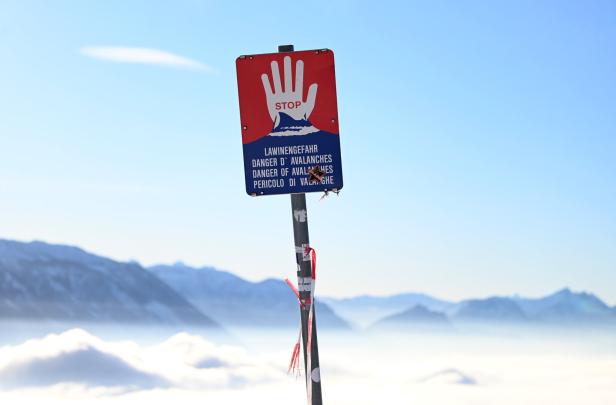 Warnschild mit roter Hand und Hinweis auf Lawinengefahr vor verschneiten Bergen und blauem Himmel.