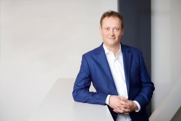 Ein Mann im blauen Anzug und weißem Hemd steht lächelnd an einer hellen Theke mit neutralem Hintergrund.