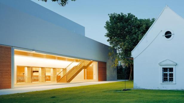 Ein moderner Bau mit großer Glasfront und Treppe steht neben einem weißen historischen Gebäude auf einer grünen Wiese.