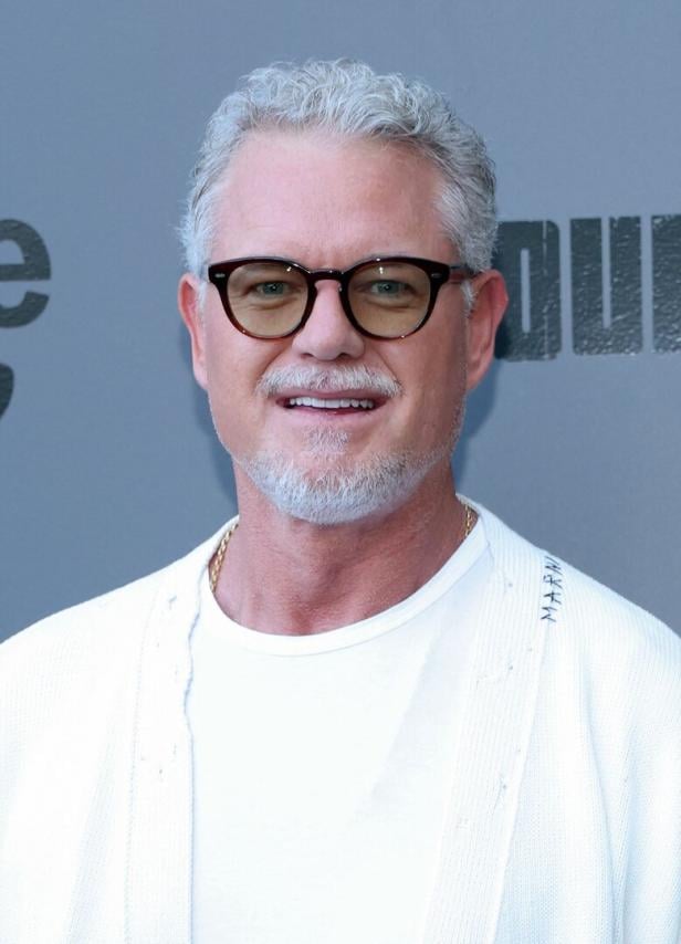 Eric Dane mit grauen Haaren und Brille lächelt in einem weißen Oberteil.