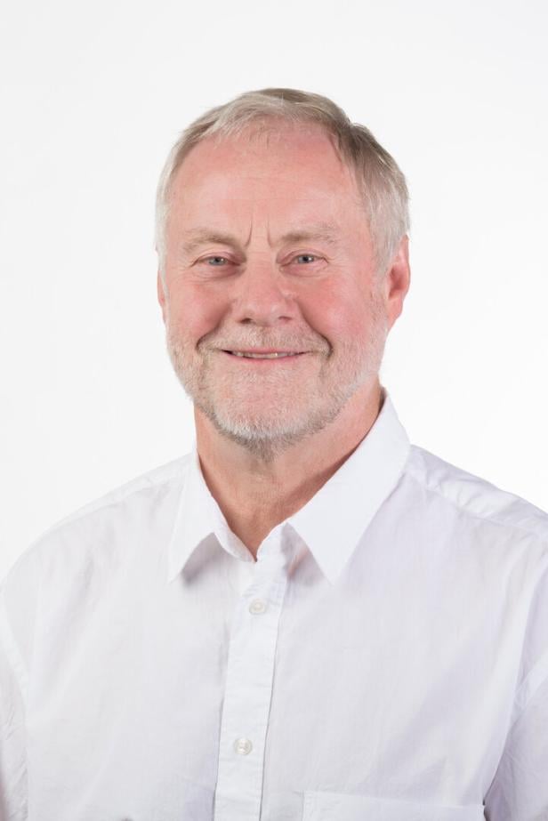 Ein lächelnder Mann mit grauem Haar und Bart trägt ein weißes Hemd vor einem weißen Hintergrund.