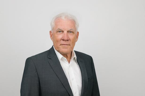 Ein älterer Mann mit grauen Haaren trägt ein dunkles Jackett und ein weißes Hemd vor einem hellen Hintergrund.