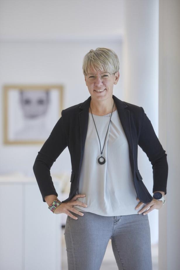 Eine Frau mit Brille, Blazer und Halskette steht mit den Händen in den Hüften da.