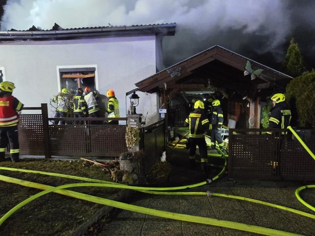 Mehrere Feuerwehrleute bekämpfen einen Brand in einem Wohnhaus, dichter Rauch steigt aus dem Dach auf.