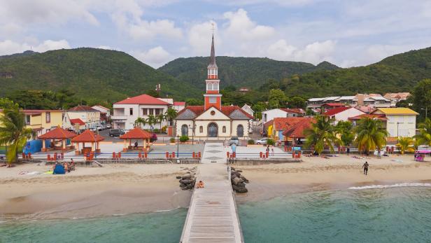Eine Kirche mit hohem Turm steht im Zentrum eines kleinen Ortes am Strand, umgeben von bunten Häusern und Palmen.