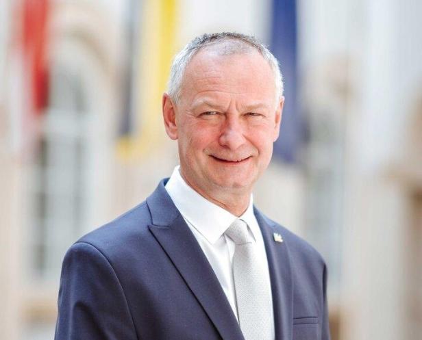 Ein lächelnder Mann mit Anzug und Krawatte steht vor einem unscharfen Hintergrund.