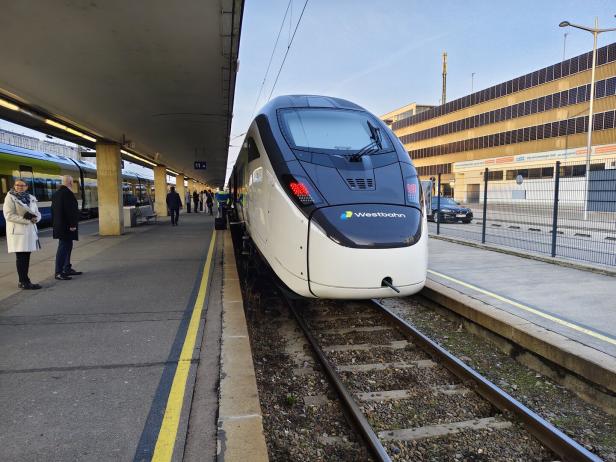 Moderner Westbahn-Zug steht am Bahnsteig, Menschen warten daneben.