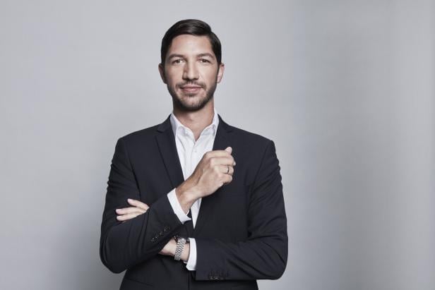 Ein Mann im dunklen Anzug mit weißem Hemd steht vor neutralem Hintergrund und blickt freundlich in die Kamera.