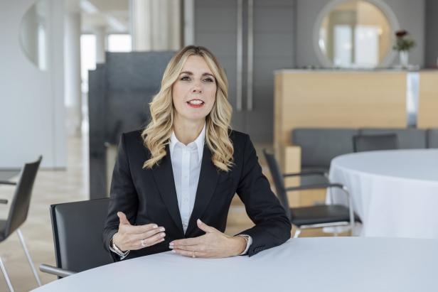Eine blonde Frau im Blazer gestikuliert an einem Tisch in einem hellen Raum.