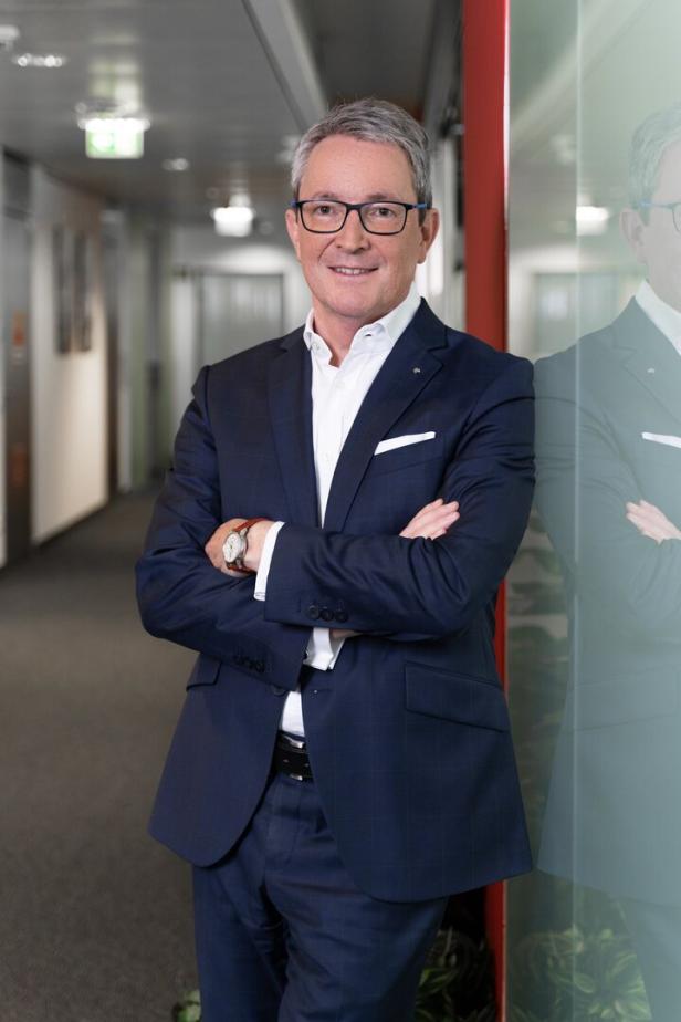 Ein Mann mit Brille und Anzug steht mit verschränkten Armen in einem Büroflur.