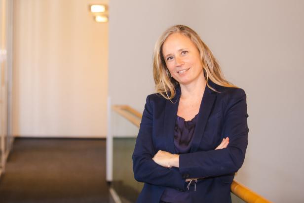 Eine Frau mit blonden Haaren lächelt, die Arme verschränkt, in einem blauen Blazer.