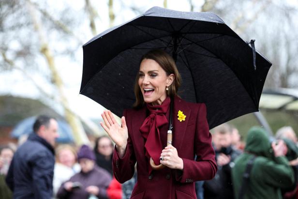 Prinzessin Kate in Wales winkt unter einem Regenschirm, gekleidet in einem bordeauxroten Mantel.