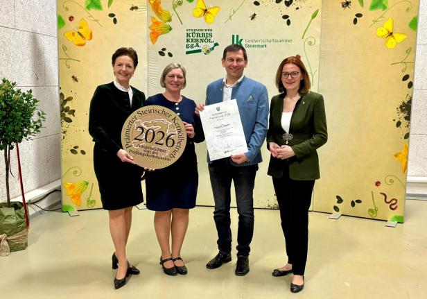 Vier Personen stehen lächelnd vor einem dekorierten Hintergrund, zwei halten eine Auszeichnung und eine Urkunde für Kürbiskernöl 2026.