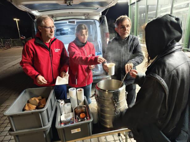Drei Personen verteilen an einem offenen Kofferraum warme Suppe und Brot an eine Person mit Kapuze.