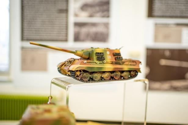 Ein detailgetreues Modell eines Tarnfarben-Panzers steht auf einem transparenten Podest in einer Ausstellung.