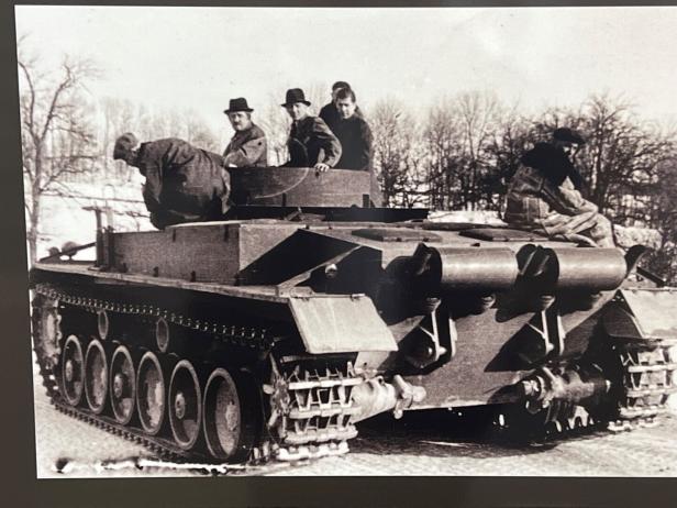 Mehrere Personen sitzen und stehen auf einem Panzerfahrzeug im Freien, umgeben von winterlicher Landschaft und kahlen Bäumen.